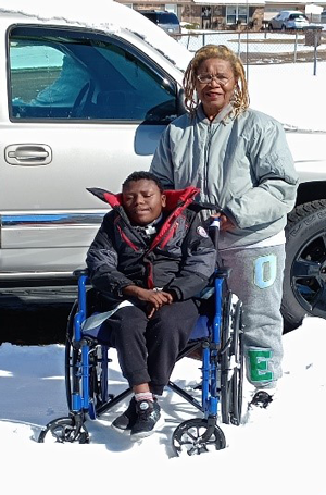 Two individuals are standing outdoors in snowy conditions. A boy is seated in a blue wheelchair and an older woman is standing behind him. A white vehicle with black rims is parked nearby, and both individuals are dressed in winter clothing, highlighting a cold environment.