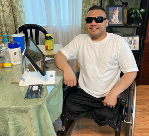 A man in a white t-shirt and sunglasses sits in a wheelchair at a table with various items in a living room.