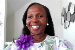 A black woman wearing a floral-patterned top with a large purple flower accessory pinned near the shoulder. Background features a white wall with a decorative arrangement of circular wooden pieces.