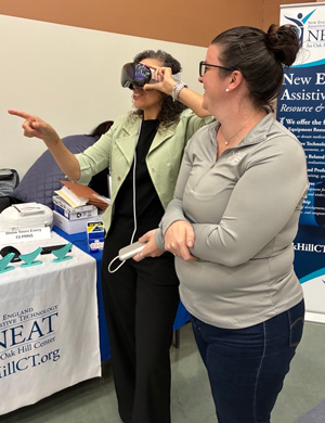 Two women at a booth; one is wearing a headset and the other is assisting her while standing next to a table with informational materials and a NEAT Center banner.