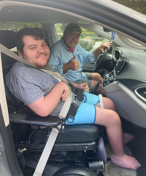 Two men seated inside a car, with one person in the driver's seat and the other in the front passenger seat using a wheelchair. Both individuals are wearing seatbelts, and the driver is giving a thumbs-up gesture