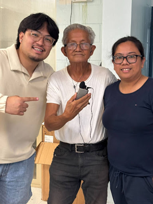 Three people standing indoors, with one person in the center holding a small device and wearing a microphone headset. The person on the left is pointing towards the center individual, while the person on the right stands close, all dressed in casual clothing.