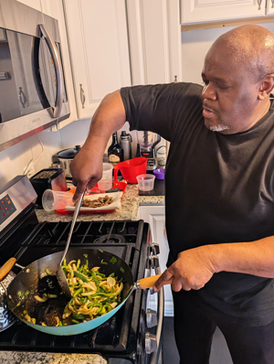 A bald Black man in a black shirt stirs vegetables in a pan on a stovetop in the kitchen