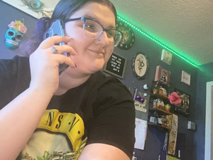 Young woman wearing glasses and a black T-shirt talking on a blue smartphone in a room decorated with dark walls and various wall hangings.