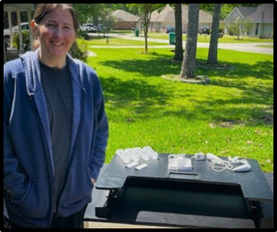 A white woman dressed in a blue jacket stands beside a table with electronics smiling at the camera.