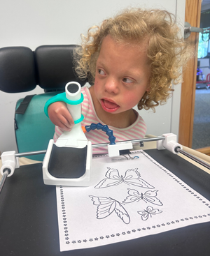 A young blonde girl with curly hair holds adapted silverware while sitting in a chair with a tray.
