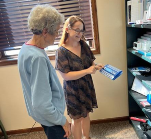 A younger woman demonstrates a device to an older woman while standing beside shelves filled with assistive technology items in an office setting.