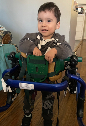 A young child using a blue and green pediatric walker designed for mobility support. The walker features padded straps, adjustable components, and a label with safety instructions.