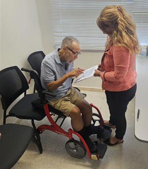 An older man in a wheelchair reviewing a document with a standing woman in an office setting. There is a desk with paperwork and office supplies in the background.