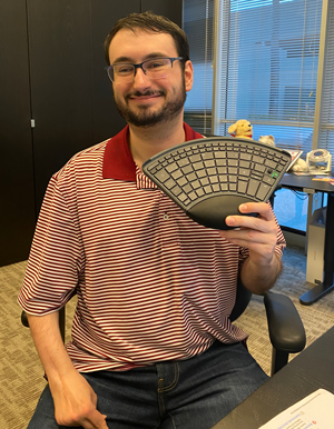 A man wearing glasses and a red striped shirt sits in an office, smiling and holding a split ergonomic keyboard.