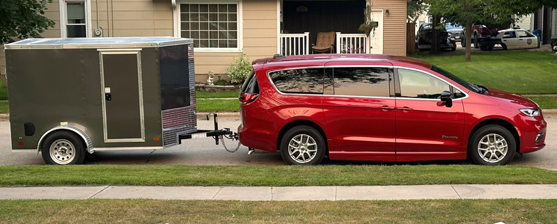 Red minivan towing a gray trailer parked on a suburban street next to a grassy lawn.