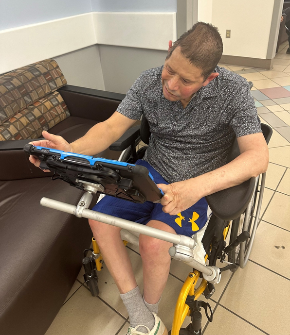 Man in a wheelchair using a tablet device mounted on a tray in a public indoor setting.