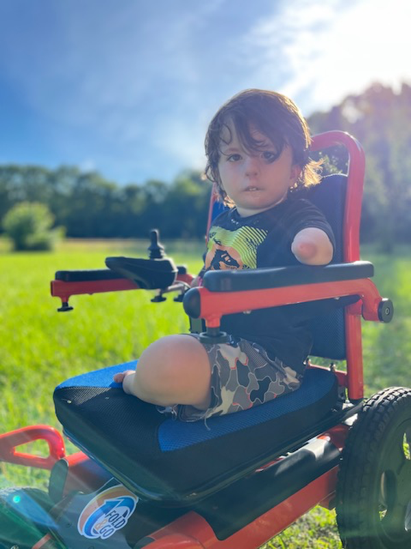 Young child sits in an adaptive wheelchair outdoors on a sunny day.