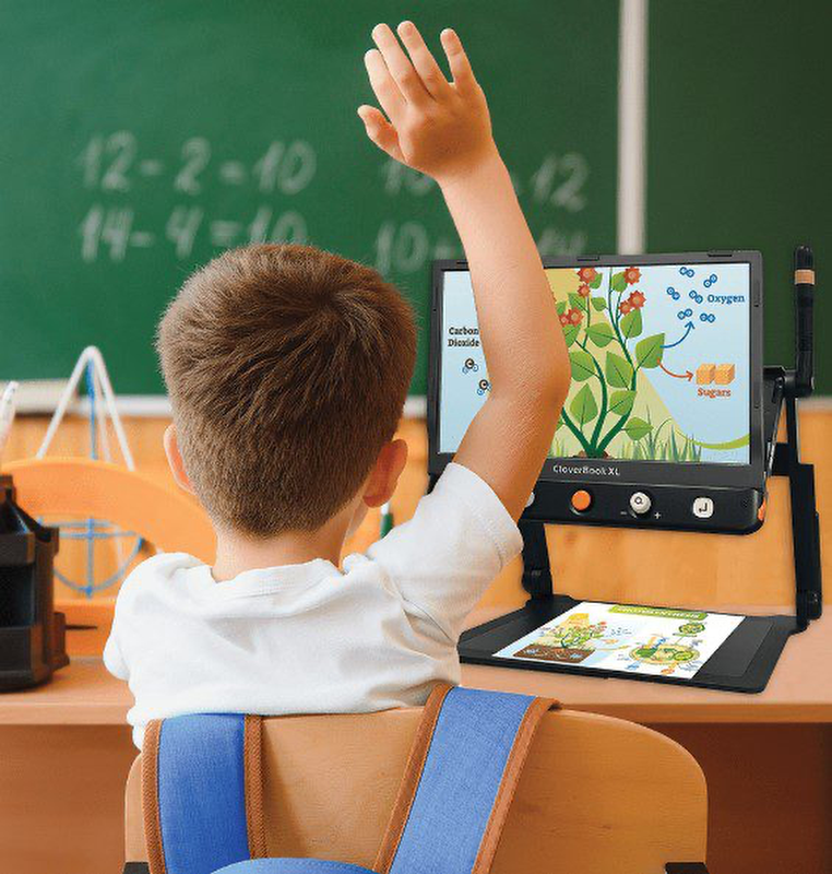 A young boy in a classroom raises his hand while using a visual aid device to view a science diagram on photosynthesis.