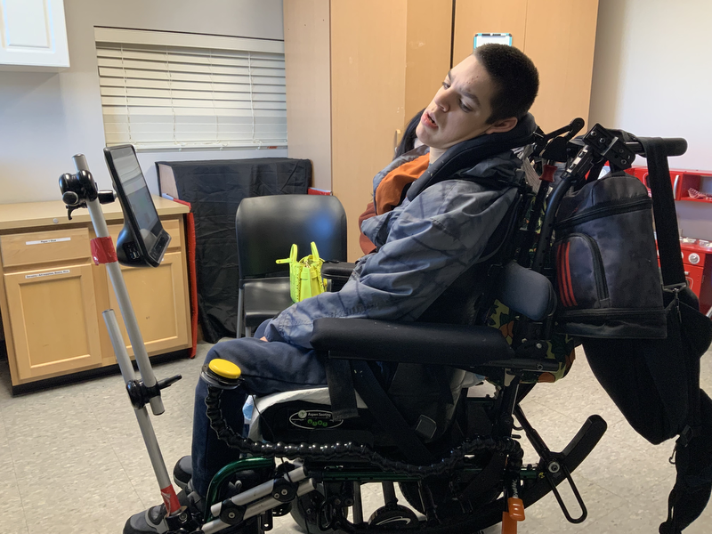A boy sitting in a wheelchair with a communication device surrounded by cabinets.