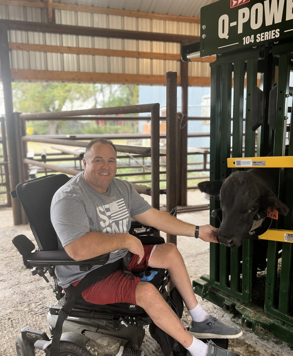 A man sitting in a power wheelchair wearing a t-shirt and shorts reaches out to touch a cow in a cattle chute.
